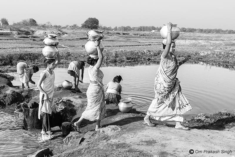 Women and a girl carry water from a traditional spring source and others wash utensils at a nearby pond in muddy paddy fields, while awaiting completion of long-delayed Jal Jeevan Mission (JJM) scheme in their Khunti district village in Jharkhand.